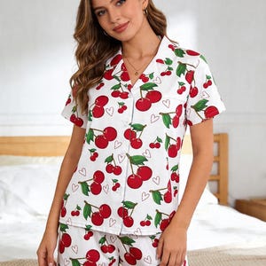 May include: White pajama set with a red cherry print. The set includes a button-down short-sleeve shirt and matching shorts. The cherries have green leaves and are interspersed with small red hearts. The pajamas are made of a lightweight fabric.