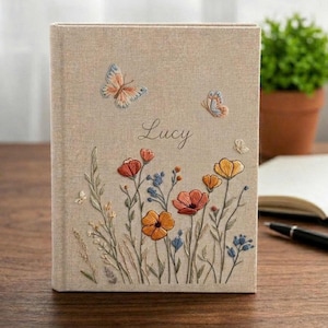May include: A beige fabric-covered book featuring embroidered wildflowers in shades of orange, yellow, and blue, along with two butterflies and the name Lucy stitched in cursive script on the front cover. The design includes various stems and leaves.