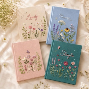 May include: Four hardback journals featuring embroidered floral designs and personalised names. The cream journal says Lindy, the blue journal says Marion, and the teal journal says Shayla. Each book displays colourful stitched wildflowers and greenery.