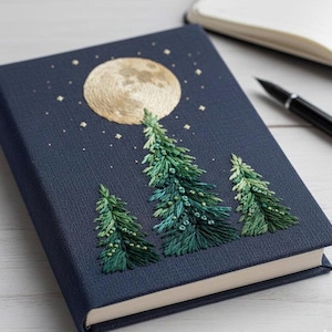 May include: A dark blue notebook featuring an embroidered design of a large, textured golden moon and three green pine trees. The night sky scene is accented with small, scattered embroidered stars in a light golden thread across the cover.
