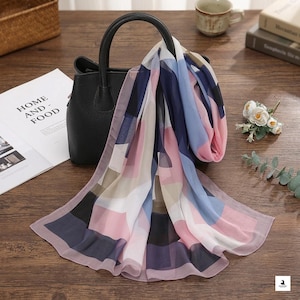 May include: A black structured handbag with two top handles, draped with a lightweight scarf featuring a geometric pattern of navy blue, light blue, soft pink, and white rectangular blocks. The scarf is made of a sheer, semi-transparent fabric.