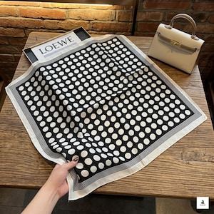 May include: A square scarf featuring a black background with a repeating pattern of white polka dots and a white border with thin black stripes. The scarf is displayed flat, showcasing its geometric design and clean, finished edges.