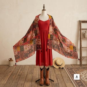 May include: A solid red midi dress with thin straps and a lace hem, paired with a wide, rectangular scarf featuring a multi-colored patchwork pattern of floral and geometric designs in shades of red, yellow, brown, and black with small black tassels.