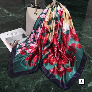 May include: A square satin scarf featuring a vibrant floral pattern with deep red, pink, and cream blossoms set against a dark emerald green background with a solid dark purple border, draped over a cream-colored handbag and a white paper card.