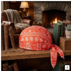 May include: A red bandana skull cap with a white paisley design, including stars, hearts, and snowflakes. The cap is tied at the back and rests on a wooden shelf with books and eyeglasses.