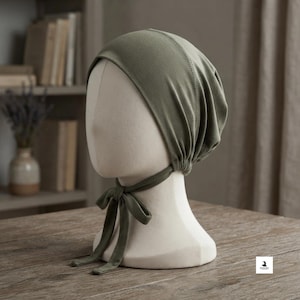 May include: An olive green head covering made of soft fabric, featuring a rounded cap design that fits snugly over the head. The accessory includes two long fabric ties at the base that can be knotted under the chin for a secure and adjustable fit.