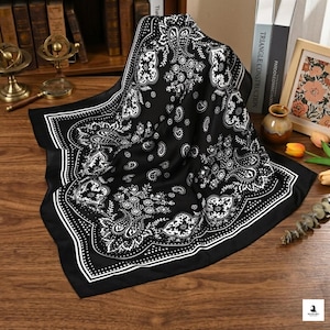 May include: Black and white paisley patterned square scarf. The scarf features a detailed design with floral and teardrop motifs. The border is a solid white line with a dotted inner line. The scarf is folded on a wooden surface.