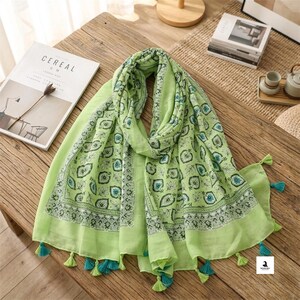 May include: A light green scarf featuring a repeating dark green and teal leaf pattern with a decorative border. The scarf is finished with small, dangling tassels in matching light green and teal colors along the edges of the fabric.