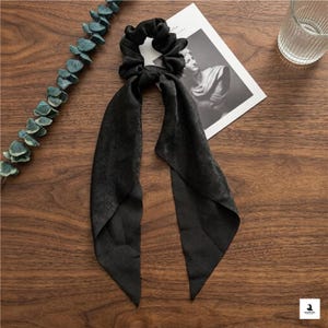 May include: Black satin scrunchie with long, flowing ribbons. The hair accessory is on a wooden surface, next to a sprig of greenery and a black and white image. A simple, elegant hair tie.