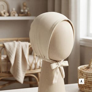 Op de afbeelding: Een effen crèmekleurig babymutsje met een ronde kroon, gestikte paneeldetails en een stoffen striksluiting onder de kin. Het mutsje wordt getoond op een gladde, neutraal gekleurde etalagepopkop, wat het eenvoudige en strakke ontwerp benadrukt.