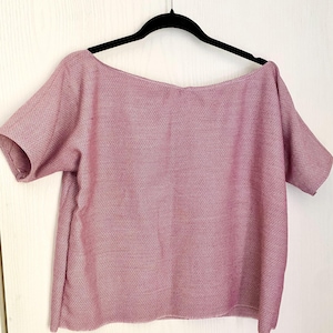 May include: A short-sleeved top in a dusty rose pink hue, featuring a wide boat neckline and a textured, woven fabric construction. The garment is displayed on a black hanger, showcasing a relaxed, boxy silhouette with simple hemmed edges.