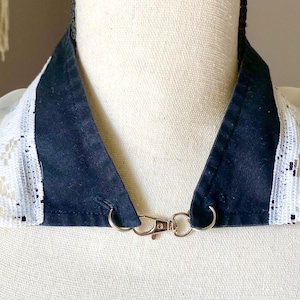 May include: A detachable collar featuring a navy blue fabric with white lace trim. The collar is secured with a silver-colored clasp. The item is designed to be worn around the neck.