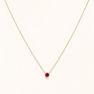 Puede incluir: Un delicado collar de oro con un pequeño colgante redondo de piedra preciosa rojo rubí. La cadena es de diseño fino y enlazado, y el colgante está centrado, creando una estética minimalista y elegante. El collar está sobre un fondo blanco liso.