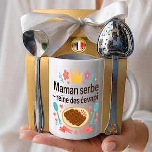 Könnte beinhalten: Ein Geschenkset mit einer weißen Tasse mit dem Text "Maman serbe - reine des ćevapi", einem herzförmigen Löffel und einem Tee-Ei. Die Tasse hat ein Kronen- und Blumenmuster. Das Set wird in einer goldenen Box mit einer weißen Schleife präsentiert.