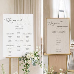 Puede incluir: Dos planos de asientos de boda con marcos y soportes dorados. Los letreros tienen un fondo claro de vetas de madera con una elegante caligrafía que dice "Take your seats" y los nombres Emma & James, y la fecha. Los arreglos florales están en la base.