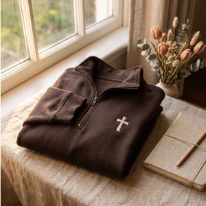 May include: A dark brown quarter-zip sweatshirt featuring a small, embroidered white cross on the left chest. The garment has a high collar, a metal zipper closure, and long sleeves with ribbed cuffs, presented in a folded, clean arrangement.
