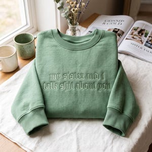 Pode incluir: Uma camisola de gola redonda em verde sálvia com o texto bordado "my sister and i talk shit about you" numa fonte gótica verde escura. A peça tem mangas compridas com punhos canelados e uma gola canelada, apresentada numa vista plana.