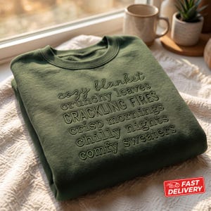 May include: An olive green crewneck sweatshirt featuring embossed text that reads: cozy blanket, crunchy leaves, CRACKLING FIRES, crisp mornings, chilly nights, comfy sweaters. The garment has long sleeves and a ribbed collar, cuffs, and hem.