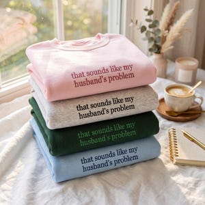 Könnte beinhalten: Ein Stapel aus vier Sweatshirts mit Rundhalsausschnitt in Hellrosa, meliertem Grau, Dunkelgrün und Hellblau. Jedes Sweatshirt ist auf der Brust mit dem gestickten Text "that sounds like my husband's problem" in Kontrastfarbe versehen.
