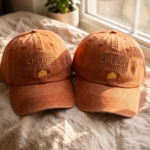 Op de afbeelding: Twee roestkleurige baseballpetten met geborduurde tekst die luidt I JUST WANT SOME CHEESE AND QUIET met een klein geel kaasblokje-icoon onder de tekst. De petten hebben een gestructureerde kroon, een gebogen klep en gestikte oogjes.