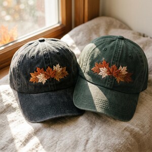 Pode incluir: Dois bonés de beisebol, um em cinza carvão e outro em verde floresta, cada um com um design bordado de várias folhas de bordo em tons de laranja, castanho e creme nos painéis frontais. Ambos os bonés têm uma aba curva e ilhós.