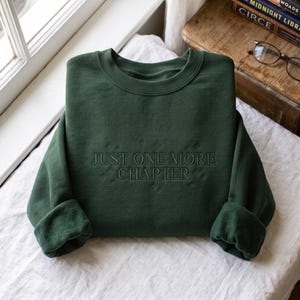 Pode incluir: Uma camisola de gola redonda em verde floresta escuro com o texto bordado "JUST ONE MORE CHAPTER" numa cor de fio a condizer, acentuada por pequenos motivos de estrelas bordados que rodeiam as letras na parte frontal da peça.