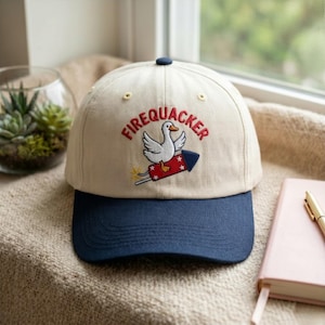 May include: A cream-colored baseball cap with a dark blue brim. The front features an embroidered graphic of a white duck riding a red firework rocket with the text FIREQUACKER arched above in red thread. The cap has small yellow eyelets.