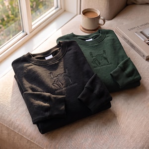 May include: A black sweatshirt and a dark green sweatshirt, both featuring a crew neckline, long sleeves with ribbed cuffs, and an embroidered graphic of a stylized creature above the text "AMAZE-AMAZE-AMAZE" centered on the chest.