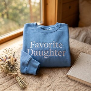 Puede incluir: Una sudadera de cuello redondo en color azul claro con el texto Favorite Daughter bordado en blanco en el centro del pecho. La prenda tiene mangas largas con pu&ntilde;os acanalados y cuello acanalado, presentada en una posici&oacute;n plegada.