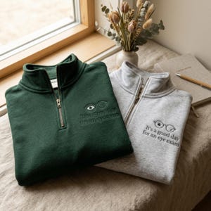 May include: A dark green and a light gray quarter-zip sweatshirt, each featuring an embroidered graphic of eyeglasses with an eye inside one lens and the text "It's a good day for an eye exam" stitched in dark thread on the upper left chest area.