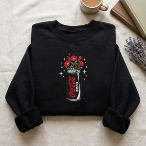 Może przedstawiać: Czarna bluza z długim rękawem z wyhaftowaną grafiką czarnej puszki napoju z etykietą Coca-Cola zero. Puszka służy jako wazon na czerwone kwiaty z zielonymi liśćmi, otoczone małymi białymi akcentami w kształcie gwiazdek.