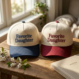 Puede incluir: Dos gorras de b&eacute;isbol con coronas color blanco hueso y viseras de colores. La gorra izquierda tiene una visera azul marino y texto bordado en azul marino que dice Favorite Daughter. La derecha tiene una visera granate y texto bordado en granate.