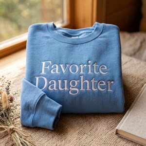 Puede incluir: Una sudadera de cuello redondo en color azul claro con el texto "Favorite Daughter" bordado en blanco centrado en el pecho. La prenda tiene mangas largas con pu&ntilde;os acanalados y un cuello acanalado, confeccionada en tela de color s&oacute;lido.