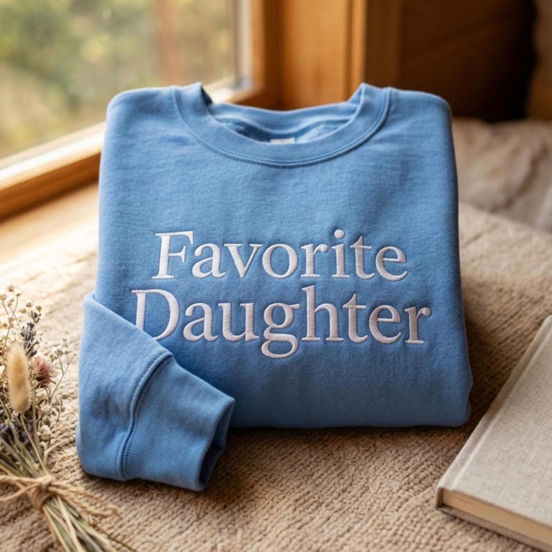 Puede incluir: Una sudadera de cuello redondo en color azul claro con el texto "Favorite Daughter" bordado en blanco centrado en el pecho. La prenda tiene mangas largas con pu&ntilde;os acanalados y un cuello acanalado, confeccionada en tela de color s&oacute;lido.