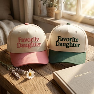 Puede incluir: Dos gorras tipo camionero con paneles frontales color crema y parte trasera de malla. La gorra izquierda tiene bordado rosa con el texto Favorite Daughter y visera rosa. La derecha tiene bordado verde oscuro Favorite Daughter y visera verde.