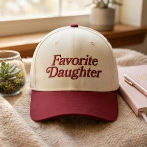 Puede incluir: Una gorra de b&eacute;isbol con la corona de color crema y la visera de color rojo oscuro. El texto "Favorite Daughter" est&aacute; bordado en rojo oscuro en la parte frontal de la corona. La gorra tiene un peque&ntilde;o bot&oacute;n rojo oscuro en la parte superior.