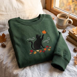 May include: A dark forest green crewneck sweatshirt featuring an embroidered design of a black cat reaching up toward a single orange maple leaf. The embroidery includes white swirling wind lines and a scattering of small red, orange, and yellow leaves.