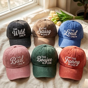 Op de afbeelding: Zes baseballpetten in zwart, bruin, blauw, roze, groen en oranje. Elke pet heeft witte geborduurde tekst: The Wild One, The Sassy One, The Loud One, The Bad One, The Boujee One en The Funny One, inclusief een klein hartsymbool.