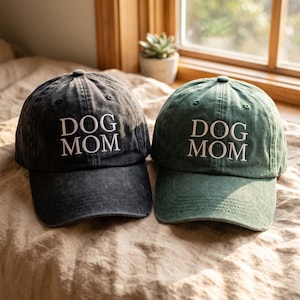 Puede incluir: Dos gorras de béisbol, una gris carbón y otra verde bosque, ambas con el texto DOG MOM bordado en letras blancas mayúsculas en la parte frontal. Cada gorra tiene una visera curva, un botón superior y un acabado de tela desgastada.