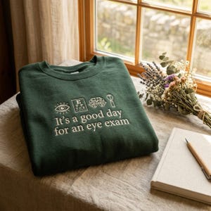 May include: A dark green crewneck sweatshirt featuring white embroidery of an eye, an eye chart, a phoropter, and an occluder, with the text "It's a good day for an eye exam" centered below the icons in a clean, sans-serif font style.