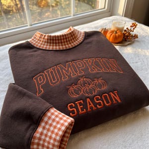 May include: A dark brown long-sleeve sweatshirt featuring a mock neck collar and sleeve cuffs in an orange and white gingham pattern. The front displays embroidered orange text reading PUMPKIN SEASON with a graphic of three pumpkins.