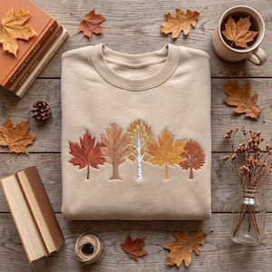 Puede incluir: Una sudadera de cuello redondo en color beige con una fila bordada de cinco árboles distintos en tonos naranja, marrón y amarillo. El diseño incluye una hoja de arce, un roble, un abedul blanco, un arce dorado y un pequeño árbol redondeado.