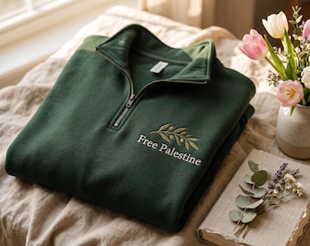 May include: A dark green quarter-zip sweatshirt featuring an embroidered olive branch design above the text "Free Palestine" in white lettering. The garment has a metal zipper pull and a folded collar, presented in a folded, flat-lay orientation.