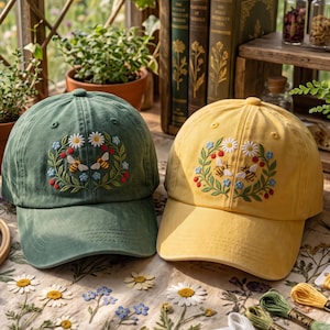 May include: A sage green baseball cap and a mustard yellow baseball cap, each featuring an embroidered design of two bees surrounded by a wreath of white daisies, blue forget-me-nots, green leaves, and small red berries on the front panel.