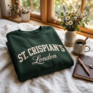 May include: A dark green crewneck sweatshirt featuring the text St. Crispian's London embroidered in a cream-colored font. The garment has a ribbed collar and long sleeves, with the text displayed in a combination of block and script lettering.