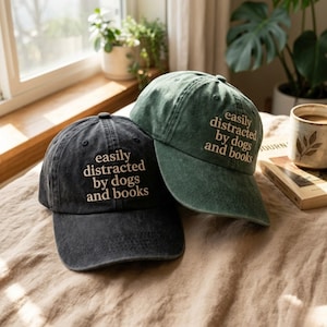 May include: Two baseball caps, one black and one green, are displayed. Both caps have the phrase "easily distracted by dogs and books" embroidered in a light tan color. The caps are sitting on a bed with a neutral-colored blanket.