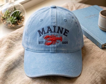 May include: A light blue baseball cap with a distressed finish, featuring dark blue embroidered text that reads MAINE and EST. 1820, centered above a detailed red lobster graphic. The cap has a curved brim and a structured six-panel crown design.