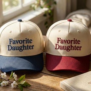 Puede incluir: Dos gorras de b&eacute;isbol con coronas de color crema y el texto bordado Favorite Daughter. Una gorra presenta texto azul marino con una visera a juego, mientras que la otra tiene texto rojo oscuro con una visera a juego.