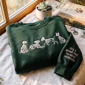 May include: A dark green crewneck sweatshirt featuring white embroidered skeleton dog designs across the chest. The sleeve includes the embroidered text: it's a good day to save animals. The garment is made of a solid-colored fabric with ribbed cuffs.