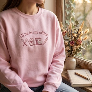 May include: A light pink crewneck sweatshirt featuring dark pink embroidery on the chest. The text reads "I'll be in my office" above icons of a whisk, an oven mitt with a heart, a stand mixer, and a rolling pin, all in a matching dark pink thread.